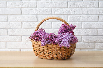 Fototapeta na wymiar Wicker basket with beautiful lilac flowers on wooden table near light brick wall