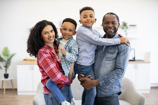 Portrait of active african american spouses holding well-behaved children in arms while feeling comfortable indoors. Loving family man and wife planning enjoyable activities with sons in apartment.