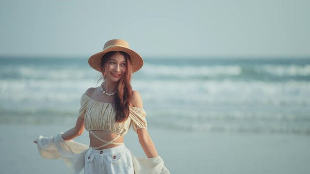 Happy Young Asian woman with straw hat walking on tropical beach, Carefree female smiling and enjoying breeze with sea in background. Travel vacation, summer outdoor pleasure.