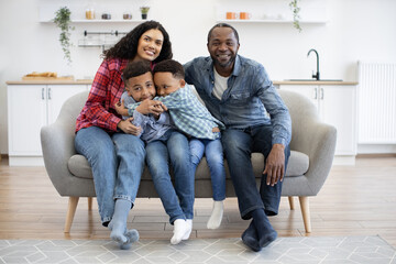 Multicultural brothers hugging each other while sitting between joyful parents on sofa indoors. Affectionate adults and kids strengthening family bonds while relaxing at cozy home.