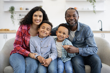 Beautiful multiethnic spouses cuddling smiling boys while making family portrait in lounge interior. Cheerful young adults showing care and affection to well-behaved sons at leisure indoors.