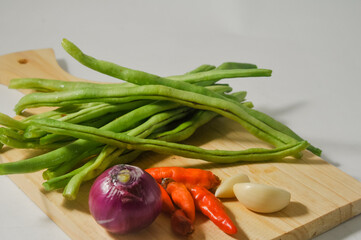 Some green beans, some red chili peppers, two cloves of garlic and a red onion on a wooden chopping board isolated on a white background