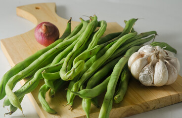 Some green beans, some red onions and a garlic bulb on a wooden chopping board isolated on a white background