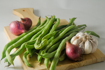 Some green beans, some red onions and a garlic bulb on a wooden chopping board isolated on a white background