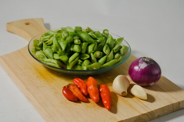 Sliced green beans on a small plate, some red chili peppers, a few cloves of garlic, and an onion on a wooden chopping board isolated on a white background