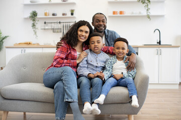 Portrait of multiracial four-person family posing together on soft couch in open-plan kitchen on sunny day. Cheerful mother and cute sons snuggling into father's arms enjoying comfort and security.
