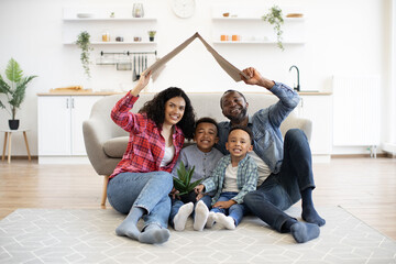 Fototapeta Multicultural family holding cardboard box above heads in form of house roof while sitting on carpet in kitchen. Smiling adults and children updating moving day checklist while resting on floor. obraz