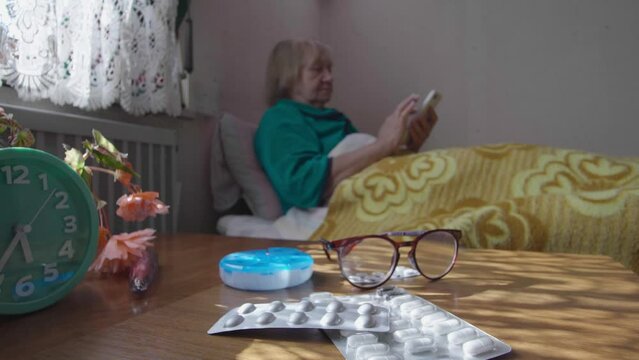 Elderly sick woman in glasses looks at the phone while lying in bed. Portrait of a sick pensioner being treated at home. Treatment concept.