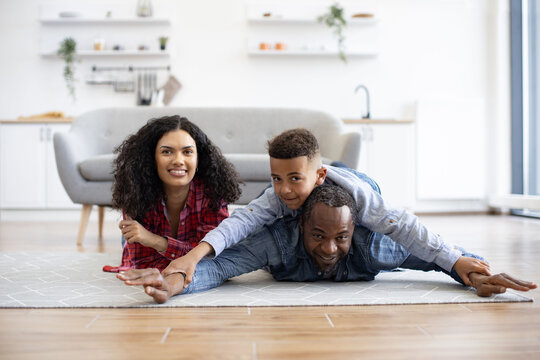 Healthy african american father holding son on back while staying with beautiful wife on kitchen floor. Affectionate dad showing swimming style to inquisitive boy while mom smiling happily aside.