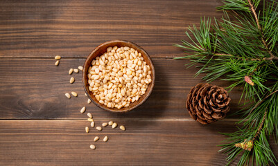 Pine nuts in a bowl on a brown wooden background with a branch of pine needles. Healthy diet snack. Top view
