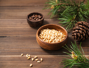 Pine nuts in a bowl and a handful of unpeeled nuts on a brown wooden background with a branch of pine needles. Healthy diet snack.