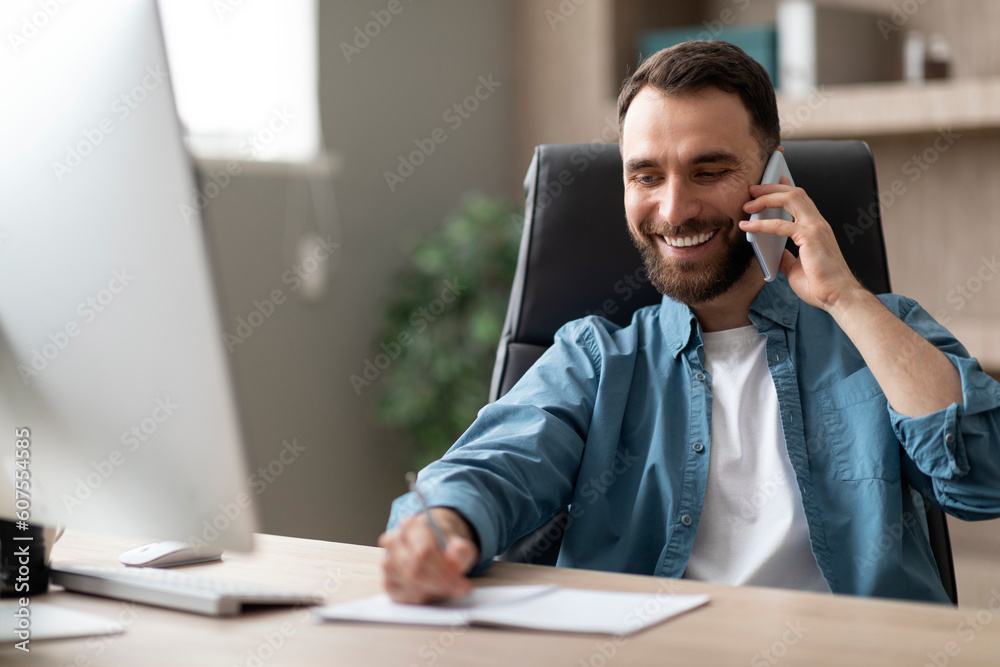 Poster smiling businessman taking notes while having phone conversation in office