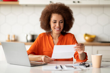 Black Woman Reading Letter While Sitting At Desk With Laptop In Kitchen