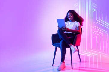 Online life. Young positive african american woman web surfing on digital tablet, sitting in armchair over neon light