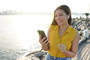 Portrait of cheerful attractive young woman wearing earphones using mobile phone outdoor with sunset in background. People and technology outdoor leisure activity.