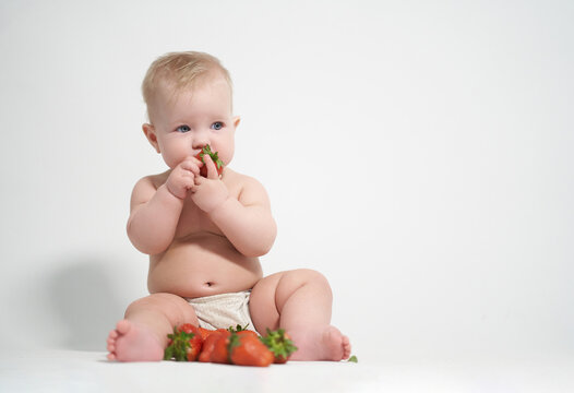 A six-month-old baby eats red strawberries, takes pictures on a light background