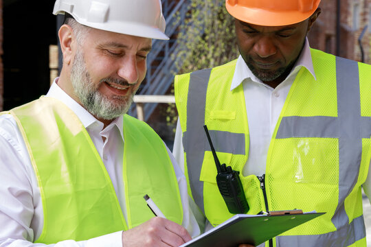 Multiracial team of competent builders in protective helmets working together on project plan outdoors. Adult caucasian foreman with clipboard recording step-by-step actions.