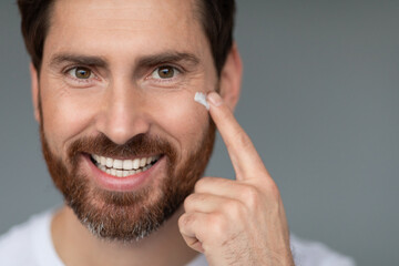 Meticulous self-care. Handsome middle aged man applying anti-aging cream under eyes, grey background, free space