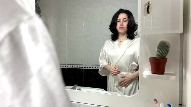 video of portrait of a young latin woman in robe in front of the mirror at home with her hands on her belly complaining of a stomach ache. pregnancies in young women are constantly increasing