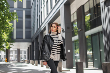 Obraz premium Beautiful fashionable vogue girl in a trendy black leather jacket with a striped pullover walks in the city