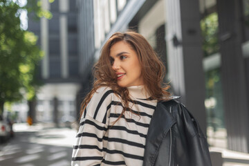 Happy beautiful curly stylish girl with a smile in a fashionable leather jacket and a striped sweater walks on a sunny day in the city