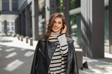 Fototapeta premium Happy cute beautiful young woman with a smile in fashionable clothes with a leather jacket and a sweater walks in the city on a sunny day