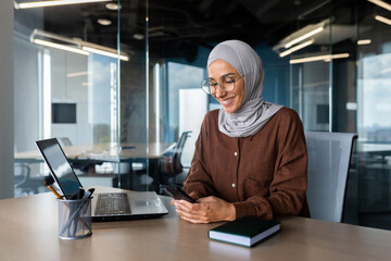 Young muslim business woman, freelancer in hijab working in office at desk and using phone smiling. Chats, types and reads messages.