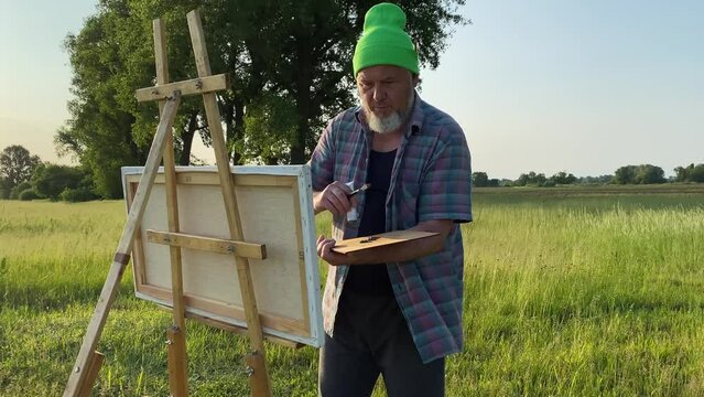 Midsection modish mature male artist oil painting landscape on canvas easel using painting tools outdoors in daybreak. Scenery view on background. Bearded middle-aged man creating artwork in daybreak