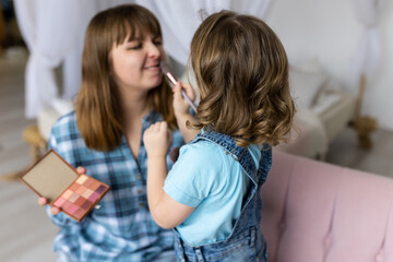 Pretty cute little girl doing make up for her mother with eye shadows palette, brush. Quality family time, enjoying domestic life, motherhood. Women's or Mother's Day celebration, having fun together