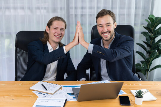 Businessmen celebrate success with joyous expressions, celebratory high-five hand gesture in office after analyzing data or achieving target. Teamwork and planning lead to successful outcome. Entity