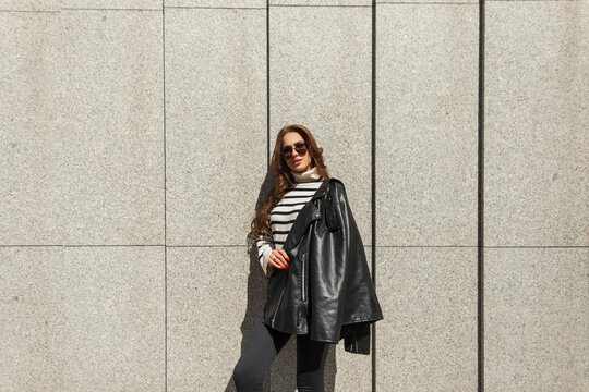 Fashionable beautiful young hipster girl with sunglasses in stylish urban rock clothes with leather jacket and striped sweater near the wall in the city