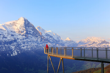 A visitor in red jacket standing on the view platform Cliff Walk at the First, Gwindelwald in the morning sunrise hours against the snow mountains