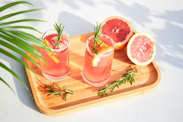 Top view of chilled summer cocktails of grapefruit or red orange, rosemary and ice on a wooden serving tray on a gray background on a bright sunny day. Vacation concept.