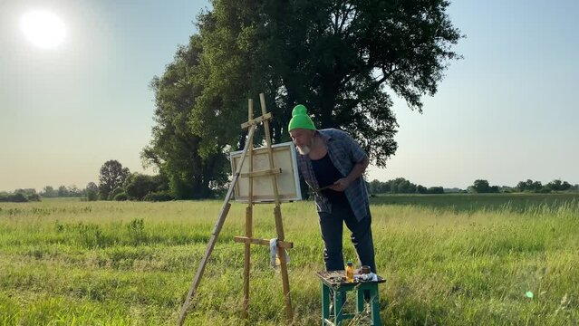 Mature bearded man artist creates oil painting examining picture by pensive look working at nature meadow in golden morning sunshine. Talented painter using palette paintbrush canvas easel outdoors