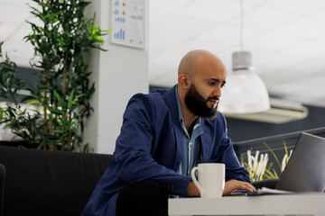 Arab marketing manager working at sales strategy on laptop in start up business office. Man entrepreneur analyzing project report while sitting on couch in coworking space