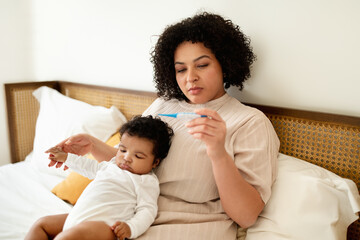 Sad worried black millennial mom with small child looks at thermometer on bed in bedroom