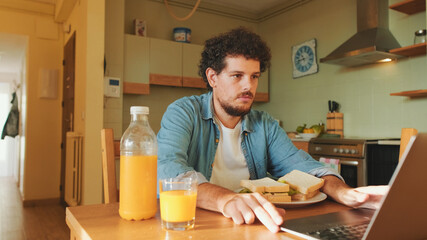 Guy dressed in denim shirt gets lunch and works on laptop while sitting in the kitchen