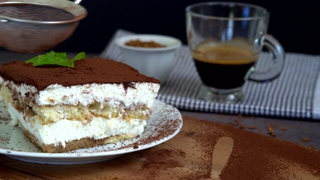Hand sprinkles cocoa powder on a traditional tiramisu cake using a sifter. Authentic Italian layered dessert with ladyfinger biscuits and mascarpone cheese. Cup of coffee in the background. Motion