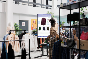 African american man looks at clothes online on touch screen monitor in fashion boutique at mall,...