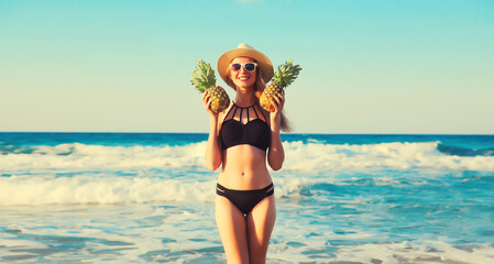 Summer vacation, beautiful happy smiling woman in bikini swimsuit and straw hat holding pineapple...