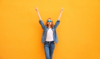 Trendy happy cheerful caucasian young woman having fun raising her hands up wearing denim jacket on yellow background