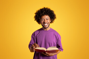 Smiling mature black curly man in purple t-shirt reading book, studying isolated on yellow...