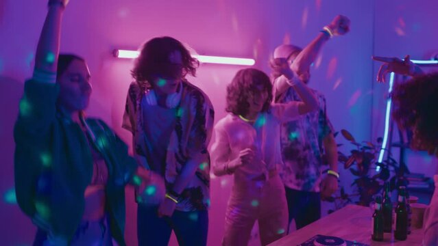 Medium shot of casually dressed young people having fun at house party, dancing and jumping around table with beer bottles and DJ turntable, in blue and pink neon disco ball lights