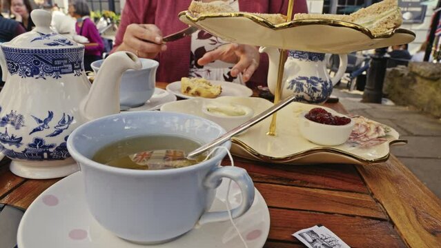 A cup of tea is steeping while a woman puts clotted cream on a scone in the background during afternoon tea on a sidewalk cafe in Windsor UK