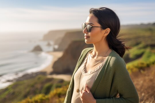 Beautiful young woman in a green sweater and sunglasses standing on a cliff with the ocean in the background
