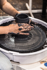 Young woman working on a pottery wheel making a cup with hands using dark black clay