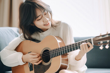 Beautiful, charming Japanese girl sitting on sofa, playing, practicing acoustic guitar in her cozy...