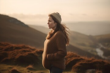 Young woman in warm sweater and hat standing on top of a hill and looking into the distance