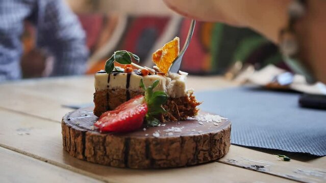 A left-handed woman takes the edge of a cake using a fork. Vegetarian cake on a wooden substrate breaks off a piece of a fork held in a woman's left hand. Close-up