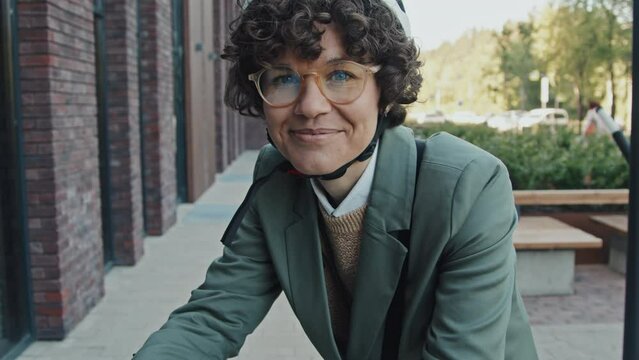 Tilt up slowmo portrait of modern Caucasian woman with curly hair wearing smart casual clothes, eyeglasses and helmet standing with bicycle smiling at camera